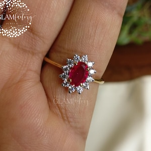 May include: A gold ring with an oval ruby centre stone, encircled by small white gemstones. The ring is displayed on a hand, with the brand name "THE GLAMfactory" visible.