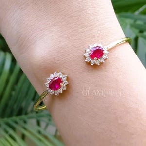 Può includere: Un bracciale a polsino dorato con due gemme ovali color rubino, circondate da piccole pietre trasparenti. Il bracciale è indossato su un polso. Il logo The Glam Factory è visibile.