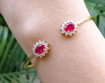 May include: A gold cuff bracelet with two oval, ruby-red gemstones encircled by small, clear stones. The bracelet is worn on a wrist. The Glam Factory logo is visible. The design is delicate and elegant.