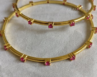 May include: Two gold bangles with pink gemstones set around the circumference. The bangles are stacked on top of each other.