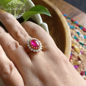 以下が含まれることがあります： 大きな楕円形の赤い宝石が、小さな透明な宝石で囲まれた金の指輪