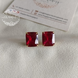 May include: A pair of square-cut, ruby-red gemstone earrings with gold-colored settings. The earrings are displayed on a white surface, with a business card in the background. The earrings are from The Glam Factory Jewels.