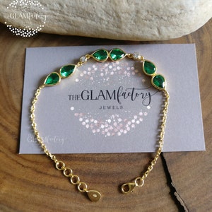 Puede incluir: Un brazalete de cadena dorada con un diseño de lágrima de piedra preciosa verde. El brazalete tiene un cierre de langosta y está adornado con pequeñas piedras de circonita cúbica transparentes. El brazalete se muestra en una tarjeta gris con el texto "THE GLAMfactory JEWELS".