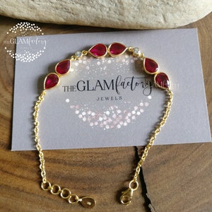 Può includere: Bracciale a catena dorata con una fila di pietre preziose rosse a forma di goccia e piccole pietre trasparenti. Il bracciale ha una chiusura a moschettone. Il bracciale è su uno sfondo grigio con il testo "THE GLAMfactory JEWELS".