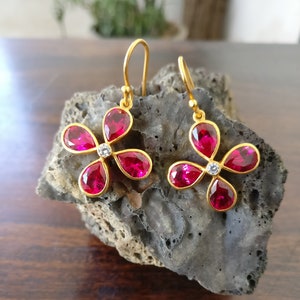 Puede incluir: Un par de pendientes de oro en forma de flor con piedras preciosas rosas y una pequeña piedra blanca en el centro de cada flor.