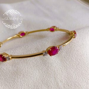 Puede incluir: Brazalete de oro con piedras preciosas de rubí ovaladas y pequeños diamantes. El brazalete tiene un acabado pulido y está sobre un fondo blanco suave. El logotipo de GlamFactory es visible.