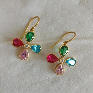 Könnte beinhalten: Ein Paar goldene Ohrringe mit einem Blumenmuster. Jeder Ohrring hat vier tropfenförmige Edelsteine in Rosa, Grün, Blau und Rot, mit einem kleinen klaren Edelstein in der Mitte.