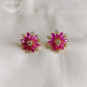 Può includere: Un paio di orecchini a lobo dorati. Ogni orecchino presenta un design floreale con petali rosa vibranti e piccole pietre di accento trasparenti. Il centro di ogni fiore ha una pietra trasparente più grande. Gli orecchini sono su sfondo bianco.