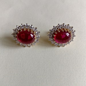 May include: A pair of gold earrings with a red gemstone surrounded by a halo of clear stones.