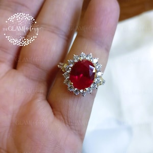May include: A gold-toned ring featuring a large, faceted, oval-shaped red gemstone, surrounded by smaller, clear stones. The ring is set on a hand, with the logo "THE GLAMfactory JEWELS" visible in the background.