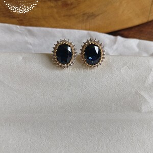 Puede incluir: Un par de pendientes de tono dorado con una gran piedra preciosa azul de forma ovalada rodeada de piedras preciosas transparentes m&aacute;s peque&ntilde;as.