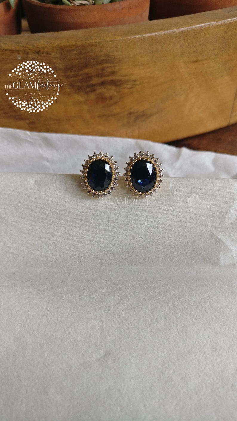 Puede incluir: Un par de pendientes de tono dorado con una gran piedra preciosa azul de forma ovalada rodeada de piedras preciosas transparentes m&aacute;s peque&ntilde;as.