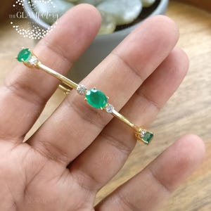 May include: A gold-toned bar brooch featuring three oval-cut green gemstones and four small round clear stones. The piece is arranged with a central green stone flanked by clear stones, with additional green stones at each end of the metal bar.