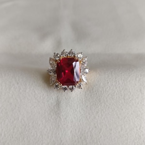 May include: A gold ring with a large, red, square-shaped gemstone surrounded by smaller, clear gemstones.