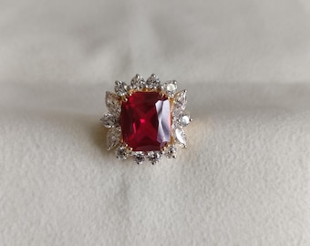Könnte beinhalten: Ein goldener Ring mit einem großen, quadratischen roten Edelstein, der von kleineren, klaren Edelsteinen umgeben ist.