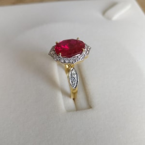 May include: A gold ring with a large red oval gemstone surrounded by a halo of smaller white gemstones.