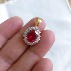 Puede incluir: Colgante de oro con una gran gema roja rubí ovalada, rodeada de pequeños cristales transparentes. El colgante tiene una argolla dorada. El logotipo de GlamFactory Jewels está en la esquina superior izquierda. Para un collar.