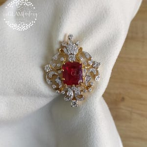 Op de afbeelding: Goudkleurige broche met een vierkante, rode edelsteen in het midden. De broche heeft een sierlijk ontwerp met heldere, sprankelende accenten en een kroonachtig element aan de bovenkant. De tekst "THE GLAMFACTORY JEWEL" is zichtbaar.