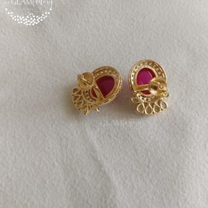 May include: A pair of gold-colored stud earrings with a ruby-red center stone. The earrings have a circular design with a halo of small, clear stones around the ruby. The Glam Factory logo is visible in the upper left corner.