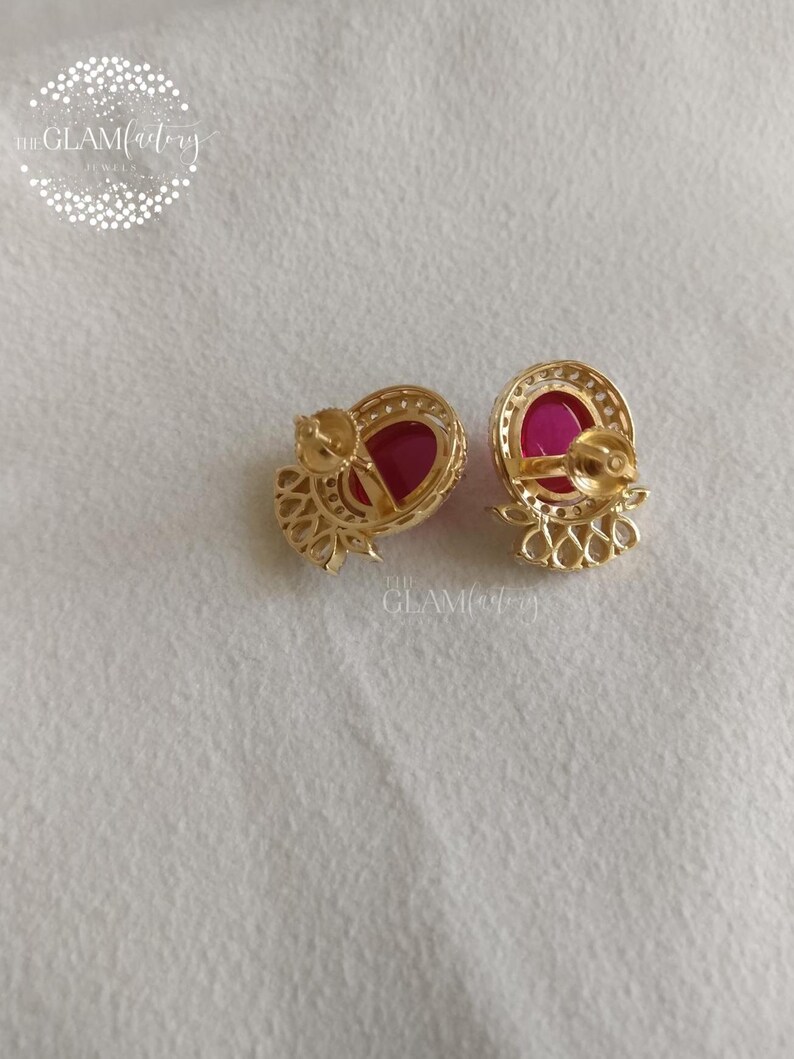 May include: A pair of gold-colored stud earrings with a ruby-red center stone. The earrings have a circular design with a halo of small, clear stones around the ruby. The Glam Factory logo is visible in the upper left corner.
