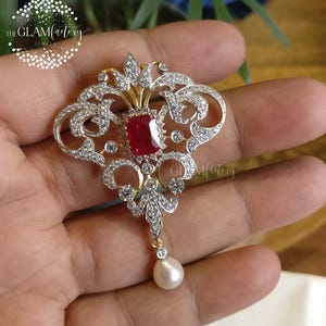 Op de afbeelding: Versierde zilveren en gouden hanger met een centrale rechthoekige rode edelsteen en talrijke kleine heldere edelstenen. Een enkele witte parel hangt van onderen. Het logo The Glam Factory is zichtbaar.