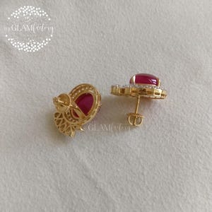 May include: A pair of gold-colored stud earrings with large, oval-shaped red gemstones. Each earring is surrounded by smaller, clear gemstones. The earrings are displayed on a white surface.