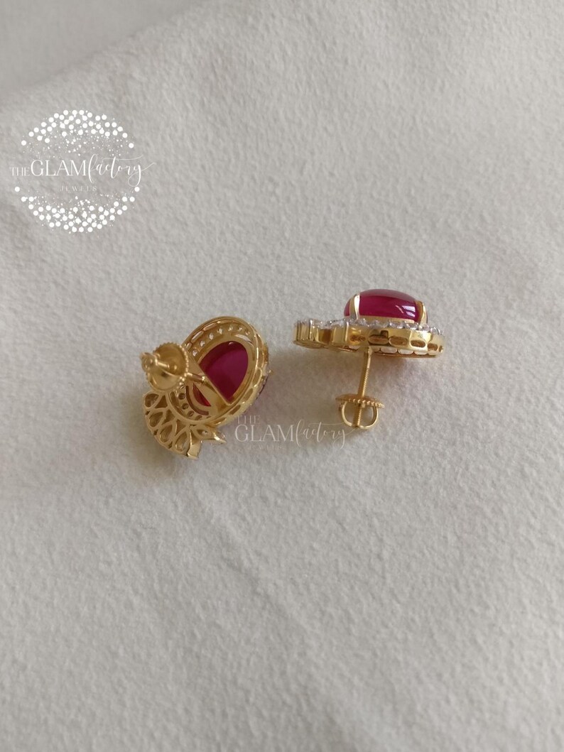 May include: A pair of gold-colored stud earrings with large, oval-shaped red gemstones. Each earring is surrounded by smaller, clear gemstones. The earrings are displayed on a white surface.