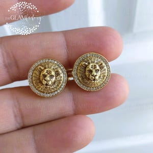 Puede incluir: Dos gemelos dorados con diseño de cabeza de león. Cada gemelo presenta una detallada cabeza de león en el centro, rodeada por un anillo de pequeños cristales transparentes. Los gemelos son redondos y tienen un acabado brillante.