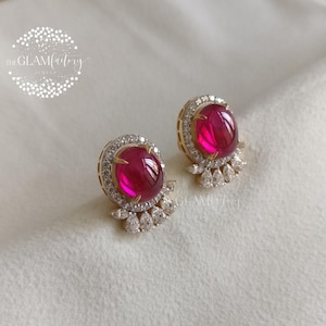 May include: A pair of gold-toned stud earrings featuring large, oval, ruby-red gemstones. Each earring is surrounded by a halo of small, clear crystals and has a cluster of crystals at the base. The Glamfactory logo is in the upper left.