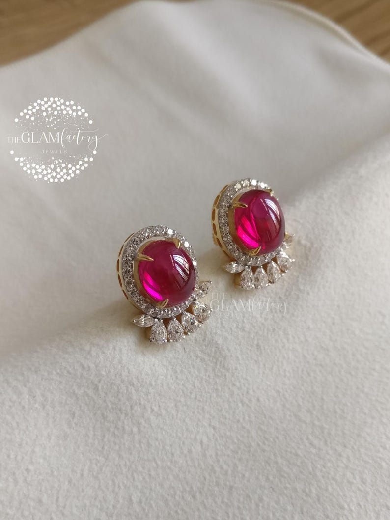 May include: A pair of gold-toned stud earrings featuring large, oval, ruby-red gemstones. Each earring is surrounded by a halo of small, clear crystals and has a cluster of crystals at the base. The Glamfactory logo is in the upper left.