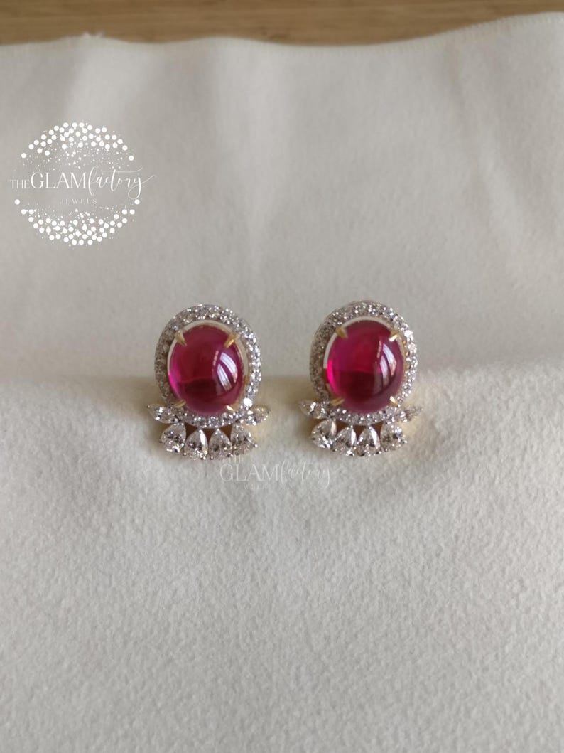 May include: A pair of stud earrings featuring large, round, ruby-red gemstones. Each earring is surrounded by a halo of small, clear crystals and set in a gold-toned metal. The earrings have a decorative, triangular crystal design at the base.