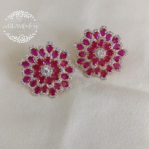 Puede incluir: Un par de pendientes de botón con forma floral. Cada pendiente presenta una piedra central redonda y transparente rodeada de piedras rojas en forma de lágrima. Las piedras rojas están rodeadas por un anillo de piedras claras más pequeñas, engastadas en un metal dorado.