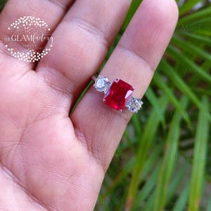 May include: A silver-toned ring featuring a large, rectangular, vibrant red centre stone flanked by two smaller, round, clear, brilliant-cut stones on a polished metal band. The design showcases a classic three-stone setting with a bright finish.