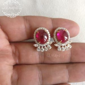 May include: A pair of stud earrings featuring oval, deep red gemstones surrounded by a halo of small, clear crystals. The earrings have a gold-colored setting and a cluster of crystals below the main stone. The Glam Factory Jewels logo is visible.