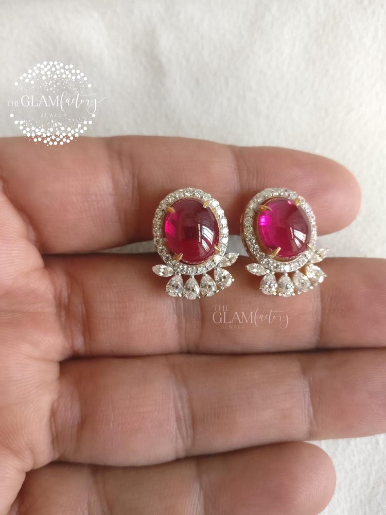 May include: A pair of stud earrings featuring oval, deep red gemstones surrounded by a halo of small, clear crystals. The earrings have a gold-colored setting and a cluster of crystals below the main stone. The Glam Factory Jewels logo is visible.