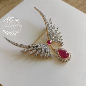 Könnte beinhalten: Gold- und silberfarbene Vogelbrosche mit einem Rubin in der Mitte und einem tropfenförmigen Rubinanhänger. Die Brosche hat ein detailliertes Design mit kleinen, funkelnden Steinen, vor einem weißen Hintergrund.