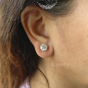 May include: A close-up of a person's ear with a large, round, clear, sparkling diamond stud earring. The earring is set in a silver setting. The text "THE GLAMfactory JEWELS" is visible in the image.