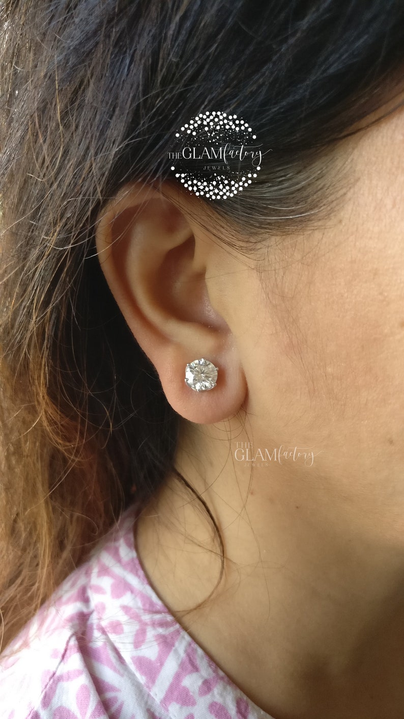 May include: A close-up of a person's ear with a large, round, clear, sparkling diamond stud earring. The earring is set in a silver setting. The text "THE GLAMfactory JEWELS" is visible in the image.
