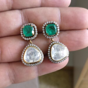 May include: A pair of gold earrings featuring a green emerald gemstone and a clear diamond-shaped gemstone. Each earring has a halo of smaller diamonds surrounding the gemstones.