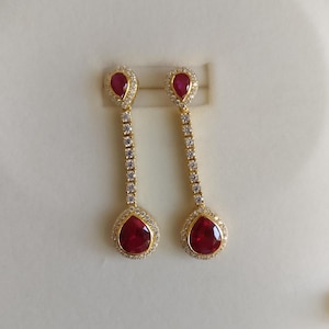 Könnte beinhalten: Ein Paar goldfarbene Ohrhänger mit birnenförmigen, tiefroten Edelsteinen. Die Ohrringe haben einen Kranz aus kleinen, klaren Steinen um die roten Edelsteine und ein lineares Design aus klaren Steinen.