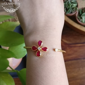 Könnte beinhalten: Goldenes Armreif-Armband mit einem roten Blumendesign. Die Blume hat fünf Blütenblätter und einen weißen Stein in der Mitte. Das Armband ist offen und hat einen kleinen weißen Stein am anderen Ende.
