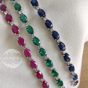 Puede incluir: Tres pulseras de piedras preciosas con piedras de rubí, esmeralda y zafiro de talla ovalada. Cada pulsera presenta una montura plateada con pequeñas piedras de acento transparentes. Las pulseras se exhiben sobre una superficie blanca.