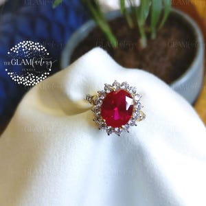 May include: A gold ring with a large, oval, bright red gemstone encircled by smaller, clear stones. The ring sits on white fabric, with a blurred background including a plant and the text "THE GLAMFACTORY JEWELS".