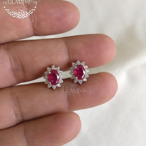 May include: A pair of ruby and diamond earrings. Each earring features a vibrant oval-cut ruby surrounded by a halo of small, sparkling diamonds. The earrings are set in a silver-toned metal and are displayed on a light background.