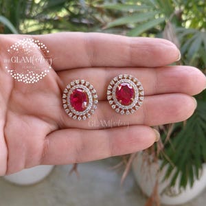 Puede incluir: Un par de pendientes de botón dorados con una piedra preciosa roja rubí ovalada central, rodeada por un halo de piedras preciosas más pequeñas y transparentes. Los pendientes son redondos. El logotipo de Glam Ladies Jewels es visible.