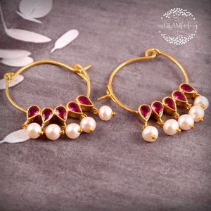 May include: A pair of gold hoop earrings with pink gemstone accents and white pearl drops. The earrings are on a grey textured surface.