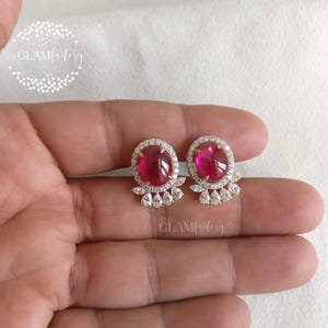 May include: A pair of stud earrings featuring oval, deep pink gemstones surrounded by a halo of small, clear stones. The earrings have a delicate, floral-inspired design at the base. The Glam Factory logo is visible.