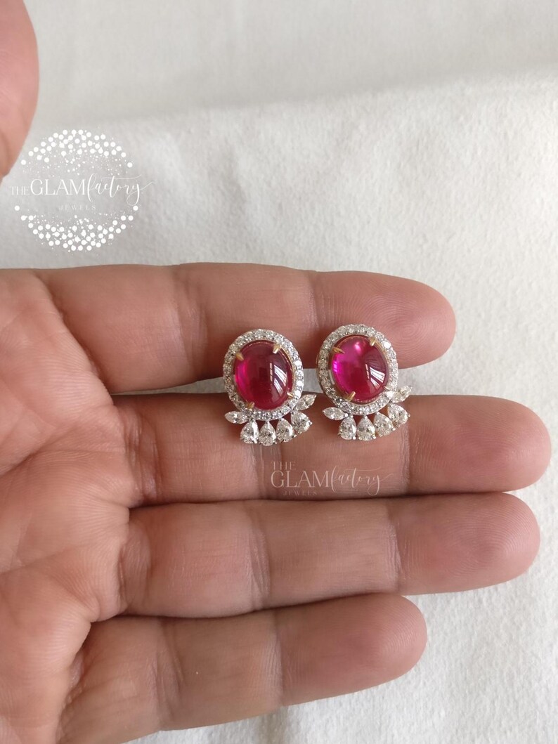 May include: A pair of stud earrings featuring oval, deep pink gemstones surrounded by a halo of small, clear stones. The earrings have a delicate, floral-inspired design at the base. The Glam Factory logo is visible.