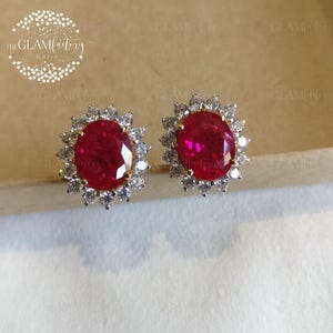 Op de afbeelding: Stud oorbellen met ovale, dieprode edelstenen, omringd door kleine, sprankelende witte stenen. De oorbellen hebben een goudkleurige zetting. Het GlamFactory-logo is zichtbaar. Op een neutrale achtergrond.