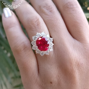 Può includere: Primo piano di un anello con una grande pietra preziosa rossa ovale, circondata da un alone di pietre preziose più piccole e trasparenti. L'anello è montato su una fascia d'argento. L'anello è su una mano con smalto d'argento.